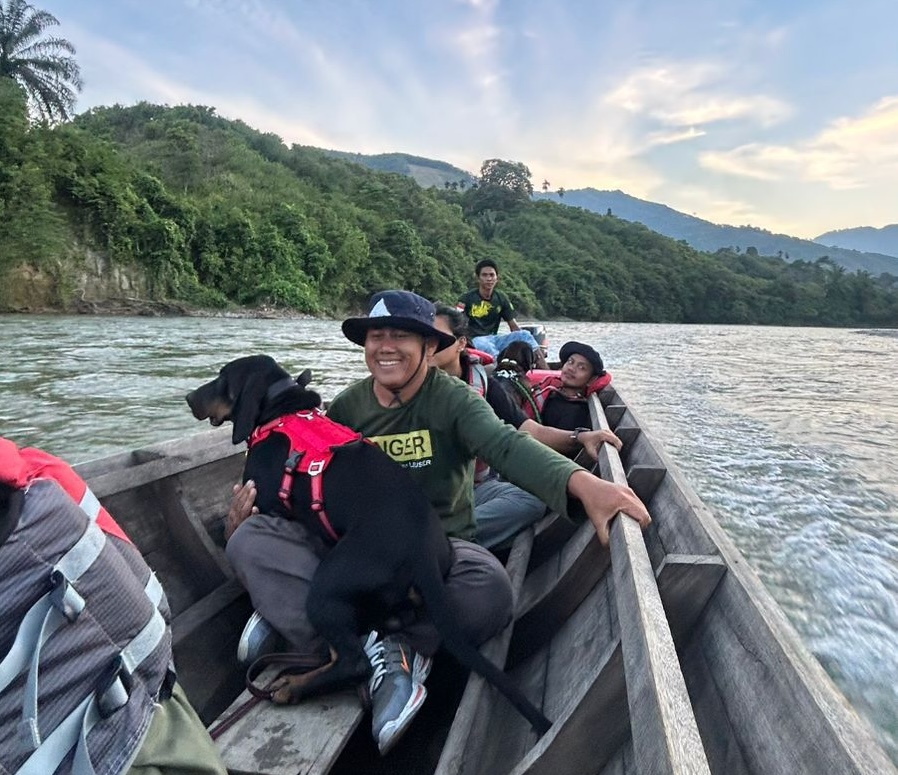 A field team travelling upriver by longboat with a detection dog, forested hills behind them